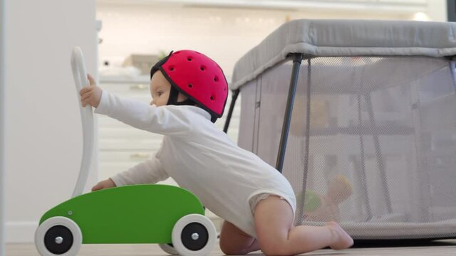 Toddler learning to stand and walk with wooden block walker, cute kid wearing baby safety helmet playing at home, small child pushing trolley around the room. High quality 4k footage