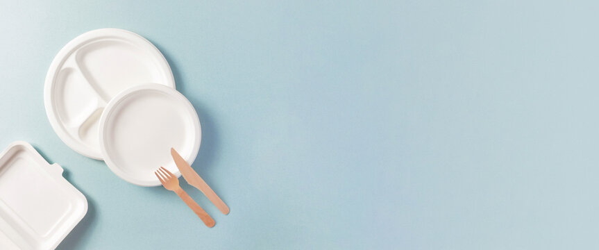 Compostable or biodegradable dinnerware flat lay composition with copy space, horizontal banner format with copy space. Paper plate, food containers, wooden fork, knife on blue. Eco-friendly concept