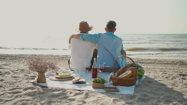 Homosexual lgbt couple, gay men having picnic at beach, sitting on blanket and embracing, looking to ech other with love