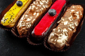 Multicolored glazed eclairs decorated with blueberries lie on a black slate plate.
