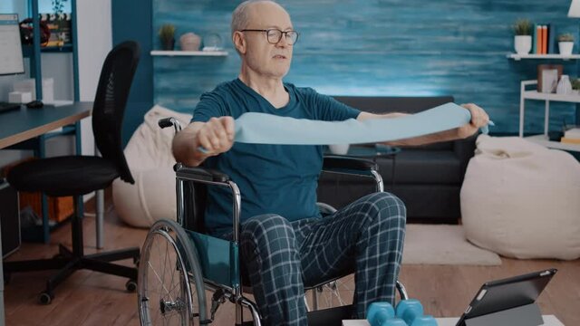Man sitting in wheelchair and doing exercise with resistance band, looking at video of workout lesson on tablet. Aged person with disability using elastic belt, watching training program