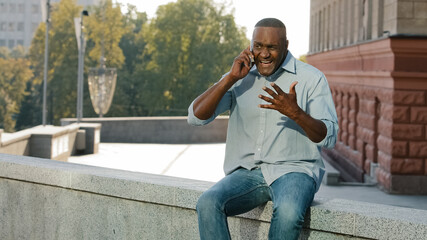 Worried nervous African American man in shirt and jeans holding mobile phone. Stressed frustrated client of retirement age talking to service sitting outdoors Customer dissatisfied with poor serve