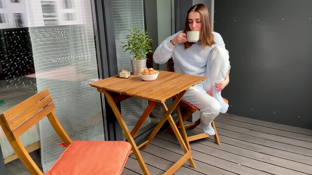 Young pretty girl is sitting on her own balcony in pajamas and drinking coffee. Modern interior.