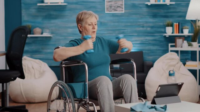 Aged woman stretching arms muscles with dumbbells, sitting in wheelchair and watching video of workout lesson on tablet. Senior person with physical disability using weights to exercise.