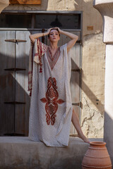 red-haired beautiful girl in Arabic clothes stands against the background of the old arabic city, bazaar. dressed in a beige abaya with embroidery and a scarf on her head