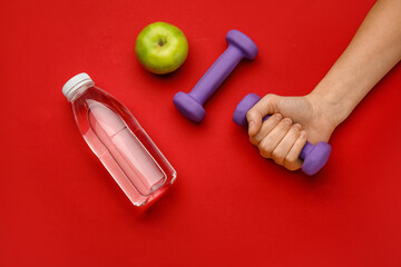 Female hand with dumbbells, apple and bottle of water on red background