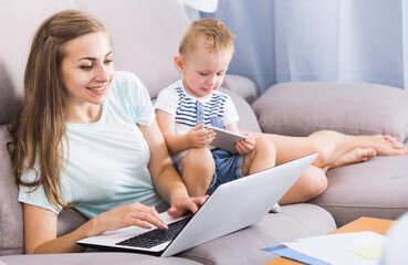 Smiling woman is productively working behind laptop while child playing on tablet at home.