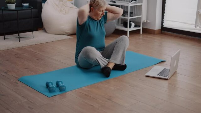Old person looking at video with workout lesson on laptop and doing physical exercise from home. Elder woman watching coach training online to practice fitness activity and sport program