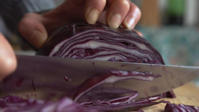 Extreme close up woman's hand slicing part of purple red cabbage with a sharp knife in the kitchen. Home cooking and healthy food concept