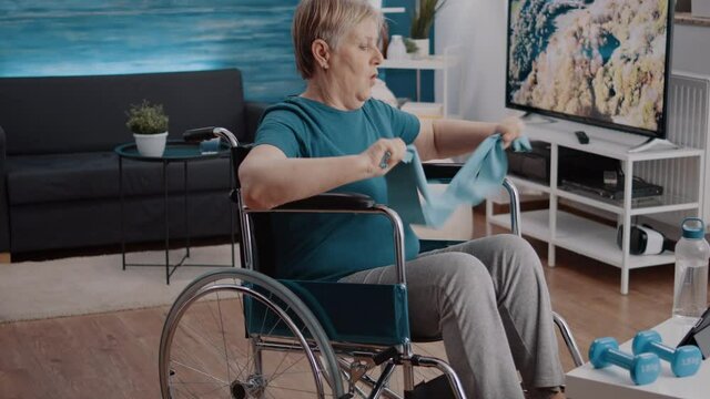 Person sitting in wheelchair and pulling resistance band to stretch arms muscles while watching workout lesson online on tablet. Old woman with disability training with elastic belt