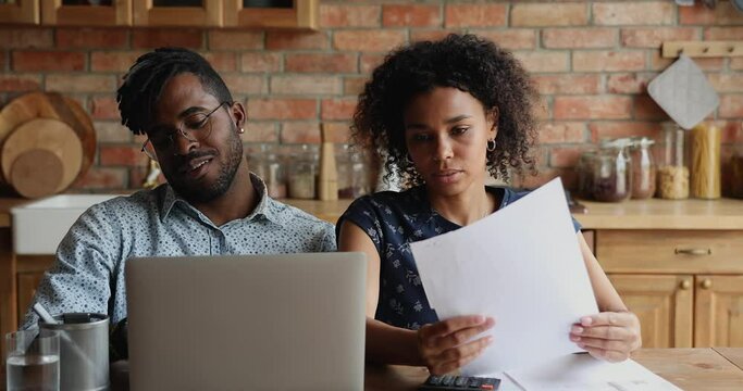 African couple sit at table with laptop, reviewing papers, using online bank app, discuss incomes smile feel satisfied, planning family budget, calculate monthly expenses, finances management concept