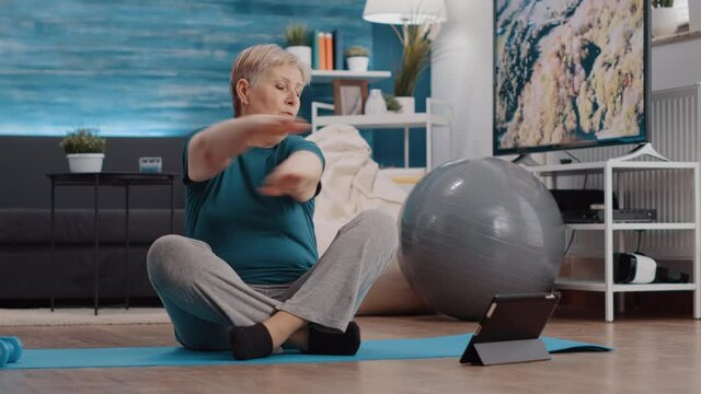Elder person doing physical exercise and watching online lesson on tablet to workout. Retired woman following training video while exercising on yoga mat. Pensioner doing sport activity