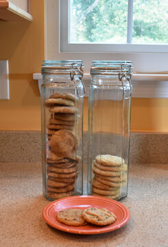 Close-up of three homemade chocolate chip cookies on an orange plate in front of two, tall, thin, glass cookie jars standing side by side