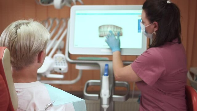 Teenager visits orthodontist. Skilled doctor in pink uniform shows jaws model spbd on computer display to teen blond boy visiting hospital office