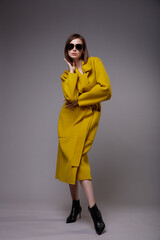 High fashion photo of a beautiful elegant young woman in a pretty yellow mustard coat, black ankle boots, stylish sunglasses posing over gray background. Slim figure. Bob haircut. Studio Shot.