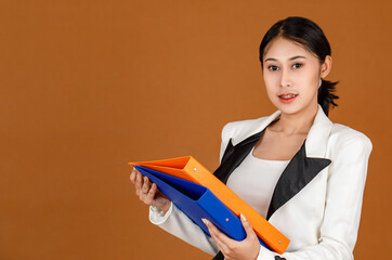Portrait closeup studio shot millennial Asian female business secretary  wears makeup in black & white fashion casual blazer standing look at camera holding hard folders in hands on brown background