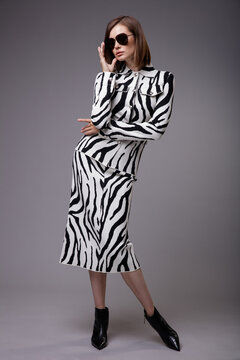 High fashion photo of a beautiful elegant young woman in a pretty zebra print suit, jacket, skirt, black ankle boots, stylish sunglasses posing over gray background. Slim figure. Bob haircut.