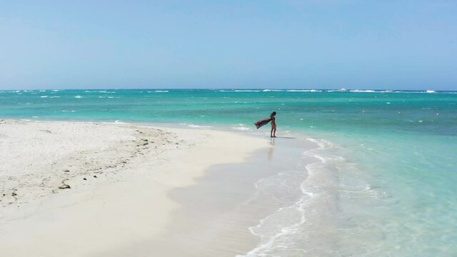 Cinematic drone shot of single woman tourist on beach, wind blowing sarong, pristine waves and water