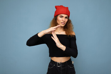 Young emotional dissatisfied pretty brunette woman wavy-haired with sincere emotions wearing black crop top and red hat isolated on blue background with free space and showing timeout gesture