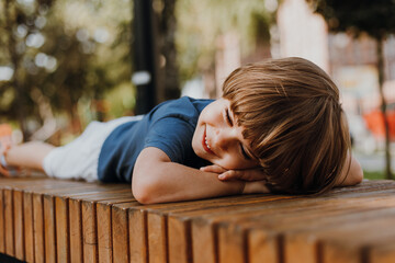 pretty little brunette boy in a blue T-shirt and white shorts is lying on a wooden bench in park....