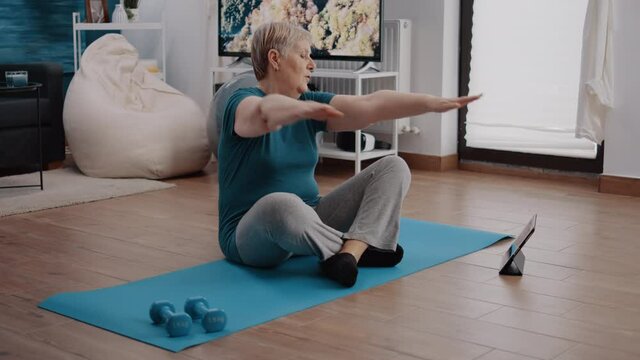 Elder woman following training lesson program on tablet while doing physical exercise and workout. Retired adult looking at gadget screen to practice aerobics and fitness activity.