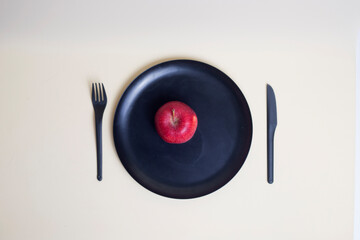 red apple on black plate with black plastic knife and fork isolated on light background flat lay