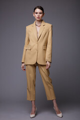 High fashion photo of a beautiful elegant young woman in a pretty  beige sand suit, jacket, pants, trousers, accessories posing on gray background. Studio Shot. Slim figure.