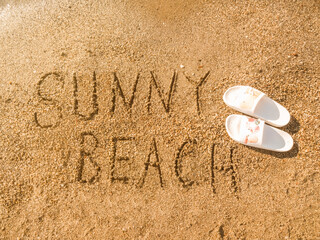 Fototapeta na wymiar The inscription Sunny beach on the sea sand and flip-flops