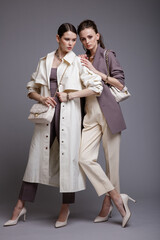 Two high fashion models in white coat,  purple jacket top, pants, accessories, handbags. Beautiful young women. Studio shot, portrait. Gray background.  Slim figure. Make up, hairstyle