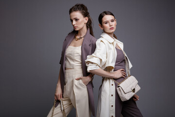 Two high fashion models in white coat,  purple jacket top, pants, accessories, handbags. Beautiful young women. Studio shot, portrait. Gray background.  Slim figure. Make up, hairstyle