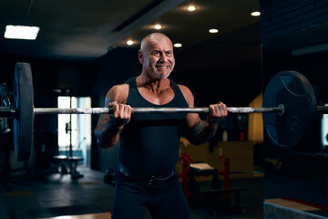 Fototapeta na wymiar Emotional Older bald Bodybuilder trains in the gym while pumping up biceps muscles with barbell