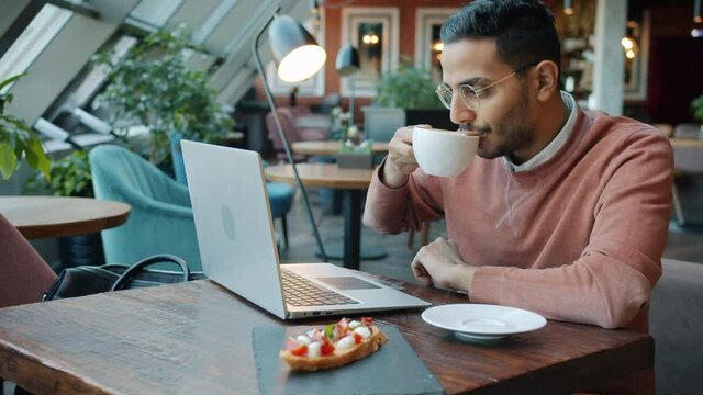 Portrait of carefree Middle Eastern student using laptop in cafe typing smiling and drinking coffee. Online communication and lunch break concept.