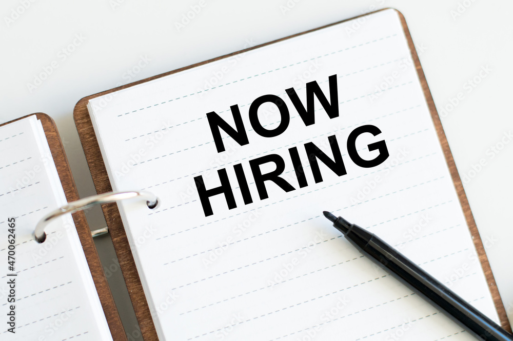 Poster text now hiring sticky on an open notebook on the table next to a black marker