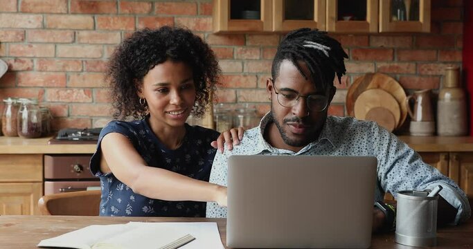 Beautiful African 30s couple sit at table in kitchen make purchase on internet using laptop, discuss order enjoy retail website, pay through e-bank app. Commerce services clients, modern tech concept