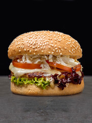 Juicy burger with grilled chicken cutlet, melted Mozzarella cheese, tomato slices, lettuce, Chinese cabbage, creamy sauce in wheat bun with sesame seeds served without plate, dark concrete background
