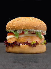 Juicy seafood burger with grilled salmon steak, melted Mozzarella cheese, tomato slices, lettuce, cabbage, bbq sauce in wheat bun with sesame seeds served without plate on concrete background
