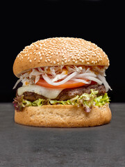 Juicy meat burger with grilled pork and beef cutlet, melted Mozzarella cheese, tomato slices, lettuce, cabbage, bbq sauce in wheat bun with sesame seeds served without plate on concrete background
