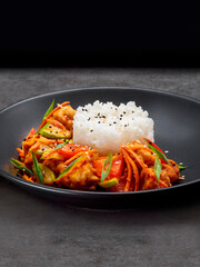 Boiled rice with chicken vegetable saute. Stewed chicken fillet (poultry), carrots, zucchini, bell pepper, green onions, served with sesame seeds on black plate isolated on background
