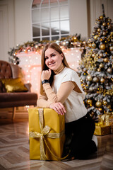 A beautiful girl in a sweater sits by a Christmas tree decorated with garlands. Next to it is a Christmas tree with boxes of gifts, decorated. New Year