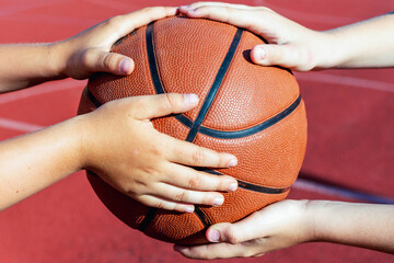 Fototapeta premium Basketball ball in the hands of a two kids,red court outdoors background.Closeup.