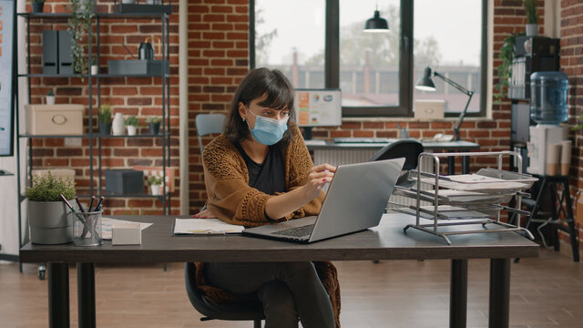 Entrepreneur checking rate charts on paper and laptop to plan business project. Woman wearing face mask and working on marketing strategy, looking at data analysis on documents.