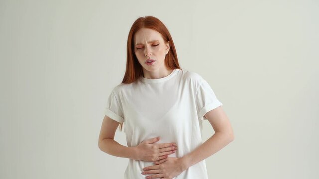 Portrait of sick young woman clasping belly and suffering from severe abdominal pain, periods spasm, irritable bowel syndrome of diet, standing on white isolated background in studio, slow motion.
