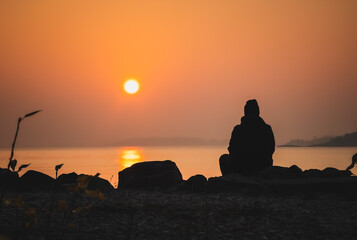 Scenic sunset by the sea. Big sun setting over the horizon. Silhouette of a man sitting alone on a rock watching the orange sundown, enjoying a peaceful moment of harmony with nature. Morning Sunrise