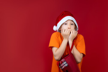 A boy wearing a Santa Claus Christmas hat on a red background clenched his fists in front of his face with fear
