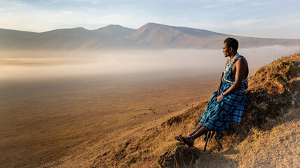 Masai Wwarrior sitting at the edge of one of Ngorongoro craters looking at the horizon and enjoying the surnrize - obrazy, fototapety, plakaty