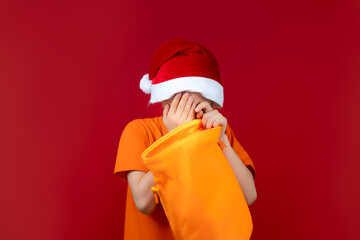 a boy in a Santa's Christmas hat on a red background holds a gift bag in his hand and is very upset