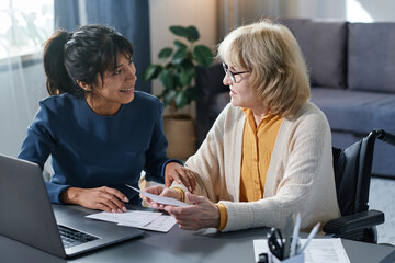 Smiling volunteer helping to senior woman to pay bills online while they sitting at the table in...