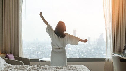 Hotel relaxation on lazy day with Asian woman waking up from good sleep on bed in weekend morning resting in comfort bedroom looking toward city view, having happy, work-life quality balance lifestyle - obrazy, fototapety, plakaty