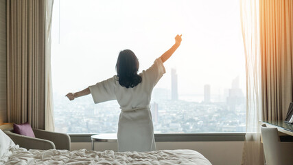Hotel relaxation on lazy day with Asian woman waking up from good sleep on bed in weekend morning resting in comfort bedroom looking toward city view, having happy, work-life quality balance lifestyle - obrazy, fototapety, plakaty