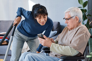 Senior man in eyeglasses using wheelchair learning to use mobile phone, he asking help to his...